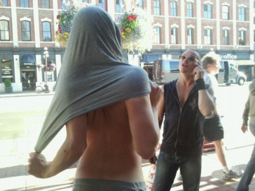 Two punks stand on sidewalk chatting while one guy puts his shirt on