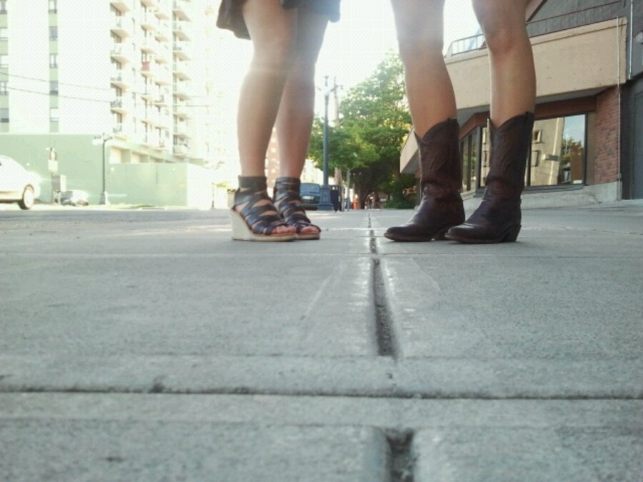 Two women stand on a sidewalk in short skirts and in cowgirl boots and shoes with straps